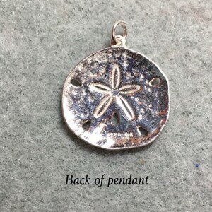 May include: A circular silver sand dollar pendant featuring a raised five-pointed star pattern and five small perforations. The metal surface has a textured, weathered appearance with the word STERLING stamped clearly on the lower front section.
