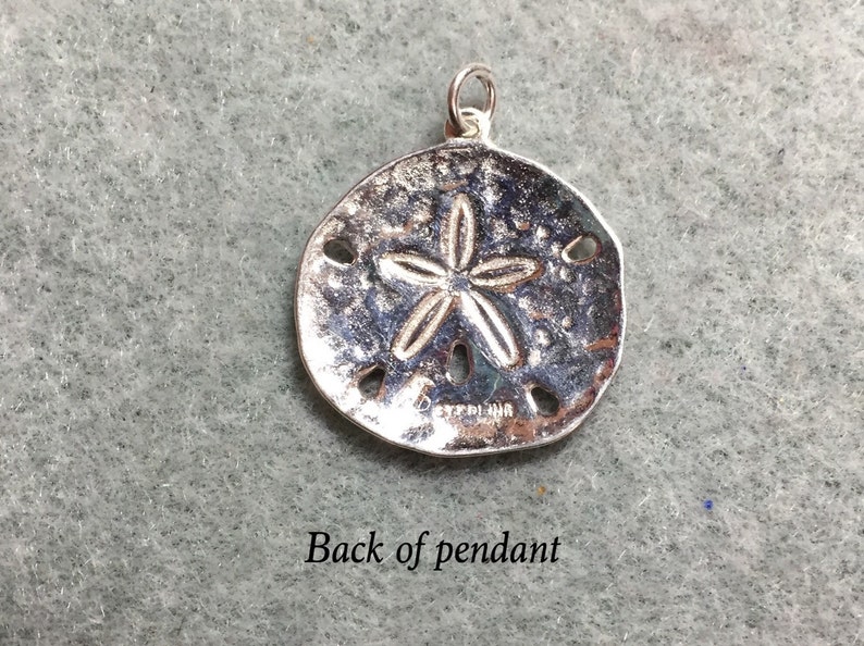 May include: A circular silver sand dollar pendant featuring a raised five-pointed star pattern and five small perforations. The metal surface has a textured, weathered appearance with the word STERLING stamped clearly on the lower front section.