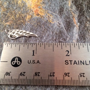 May include: A silver metal angel wing charm measuring approximately 1 inch in length. The wing is detailed with a delicate, intricate design.