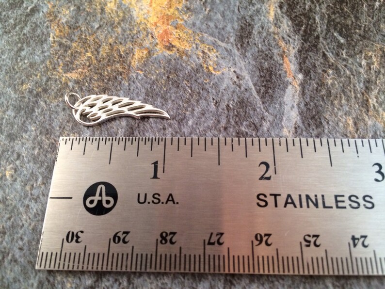 May include: A silver metal angel wing charm measuring approximately 1 inch in length. The wing is detailed with a delicate, intricate design.