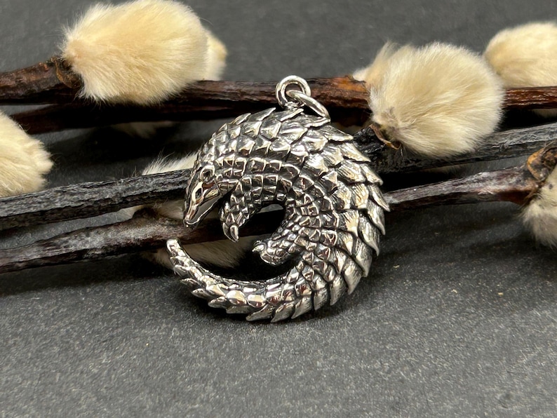 May include: A detailed silver pendant in the shape of a pangolin, featuring textured scales and a curled body. The pendant is suspended from a small loop. The background includes brown twigs and fluffy, light-colored buds, set against a dark surface.