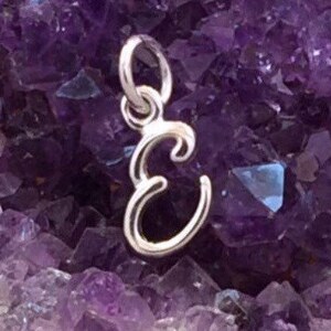 May include: A silver-colored pendant in the shape of the letter "E" hangs against a background of purple amethyst crystals. The pendant has a loop at the top for a chain.