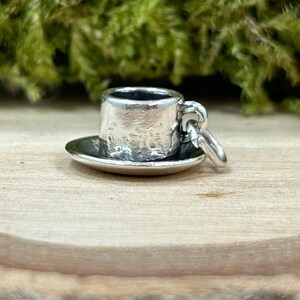 May include: A silver charm in the shape of a small teacup and saucer. The charm has a textured surface and a loop for attaching to a bracelet or necklace.