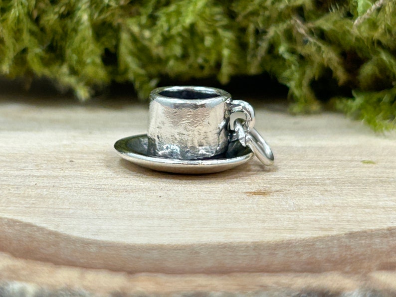 May include: A silver charm in the shape of a small teacup and saucer. The charm has a textured surface and a loop for attaching to a bracelet or necklace.