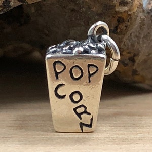 Może przedstawiać: Srebrny charm w kształcie wiaderka na popcorn z napisem "POP CORN" na boku.