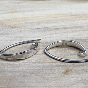 May include: A pair of silver-colored, open-hoop earrings. One earring has a hammered texture, while the other is smooth. The earrings are displayed on a light-colored wooden surface.