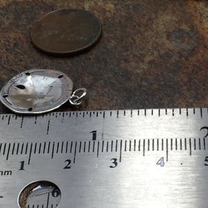 May include: A silver sand dollar charm pendant with a small jump ring attached to the side. The charm is positioned next to a metal ruler showing a diameter of approximately 0.75 inches, with a copper penny placed nearby for scale.