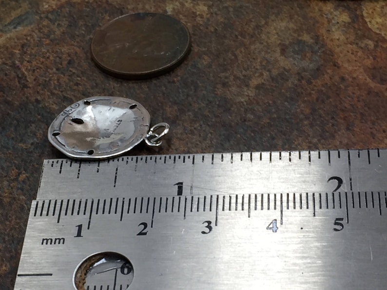 May include: A silver sand dollar charm pendant with a small jump ring attached to the side. The charm is positioned next to a metal ruler showing a diameter of approximately 0.75 inches, with a copper penny placed nearby for scale.