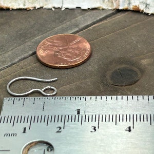 May include: A silver earring hook measuring approximately 1 inch in length. The hook is shown next to a ruler and a US penny for size comparison.