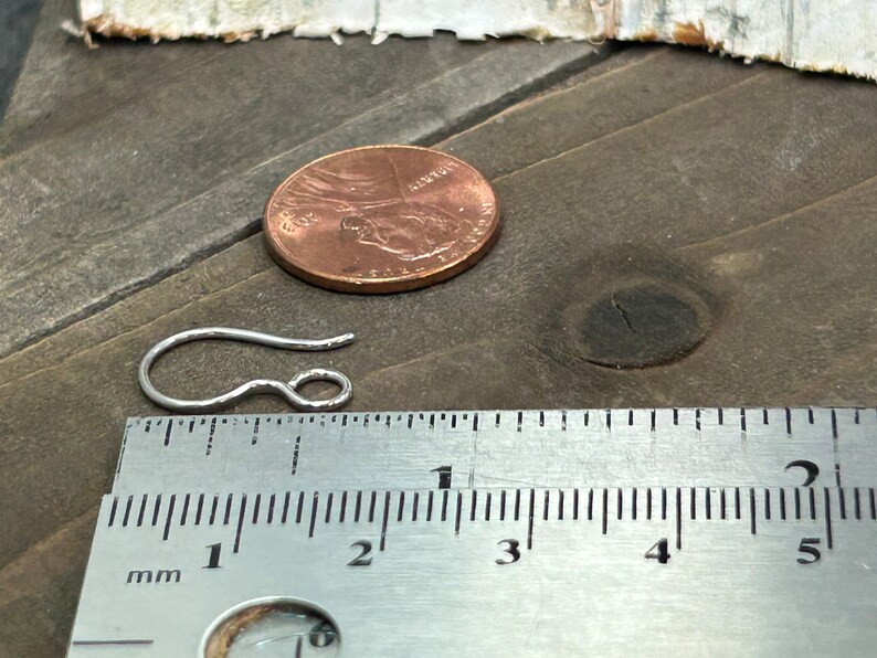 May include: A silver earring hook measuring approximately 1 inch in length. The hook is shown next to a ruler and a US penny for size comparison.