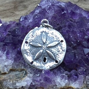 May include: A circular silver pendant featuring a textured surface with a raised five-petaled flower-like design and small perforations, topped with a small metal jump ring for attachment, resting against a cluster of deep purple amethyst crystals.