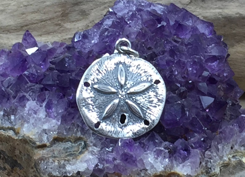 May include: A circular silver pendant featuring a textured surface with a raised five-petaled flower-like design and small perforations, topped with a small metal jump ring for attachment, resting against a cluster of deep purple amethyst crystals.
