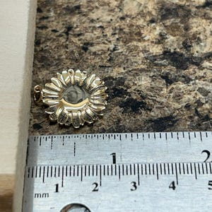 May include: A gold-colored sunflower pendant with detailed petals. The pendant is approximately 0.75 inches in diameter. The pendant is shown next to a ruler with millimeter markings.
