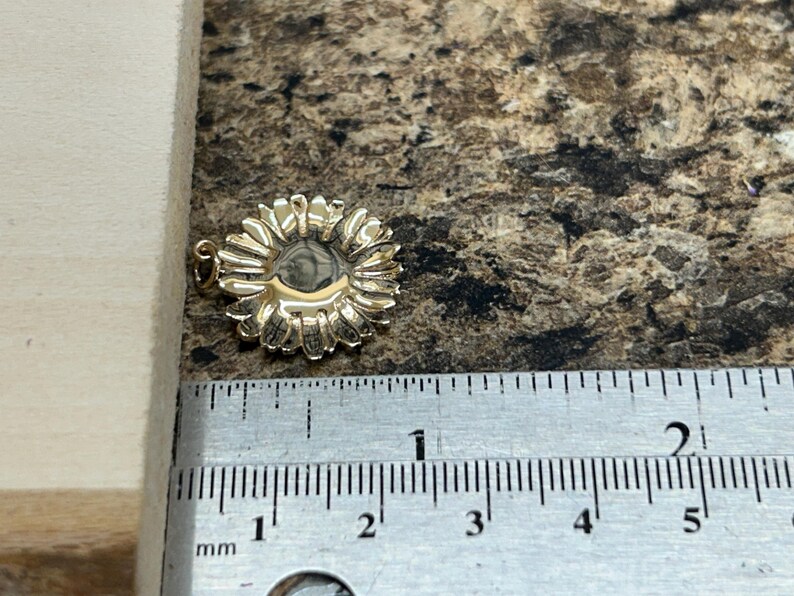 May include: A gold-colored sunflower pendant with detailed petals. The pendant is approximately 0.75 inches in diameter. The pendant is shown next to a ruler with millimeter markings.