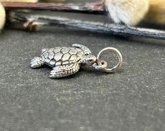 May include: A silver-colored turtle charm with a textured shell and flippers. The charm has a small loop for attaching to a necklace or bracelet. The charm is on a dark gray surface.