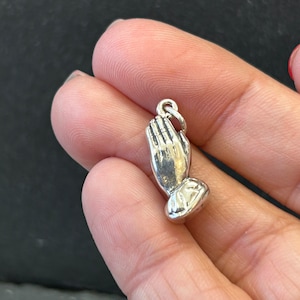 May include: A silver-colored charm in the shape of praying hands. The charm has detailed fingers and a loop at the top for attaching to a necklace or bracelet. The charm is likely made of a metal alloy.