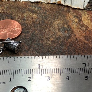 Puede incluir: Un amuleto de plata en forma de carrete de hilo con una aguja. El amuleto mide aproximadamente 2,5 cm de largo y est&aacute; al lado de una regla para referencia de tama&ntilde;o.