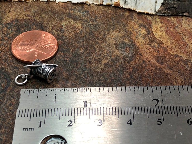 Puede incluir: Un amuleto de plata en forma de carrete de hilo con una aguja. El amuleto mide aproximadamente 2,5 cm de largo y est&aacute; al lado de una regla para referencia de tama&ntilde;o.