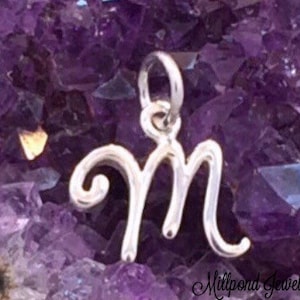 May include: A silver charm in the shape of the letter M, with a decorative swirl on the left side of the letter. The charm is photographed against a background of purple amethyst crystals.