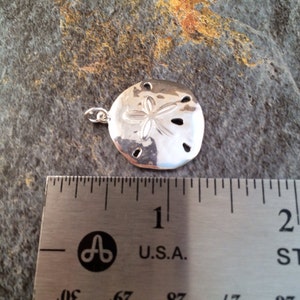 May include: A polished silver-toned sand dollar pendant featuring a central five-petal star design and five small perforations. The pendant is positioned above a stainless steel ruler, showing a width of approximately 1.25 inches.