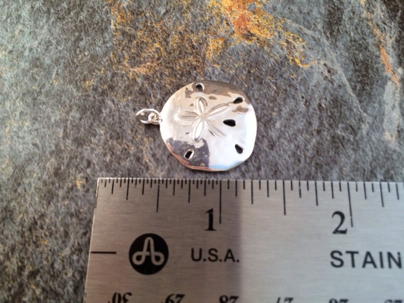 May include: A polished silver-toned sand dollar pendant featuring a central five-petal star design and five small perforations. The pendant is positioned above a stainless steel ruler, showing a width of approximately 1.25 inches.