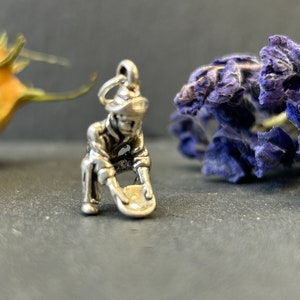 May include: A silver charm depicting a prospector panning for gold. The charm shows a figure in a hat and work clothes, bent over a pan. The charm is detailed and has a loop for attaching to a bracelet or necklace.
