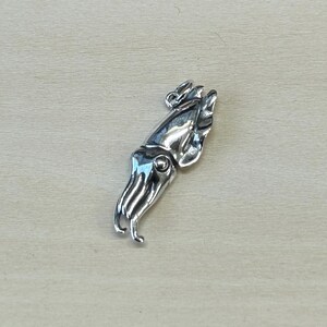 May include: A silver-colored squid pendant. The pendant features a detailed design of a squid with tentacles and a rounded body. The pendant is likely made of metal and has a loop for attaching to a chain or cord. The background is a light wood surface.