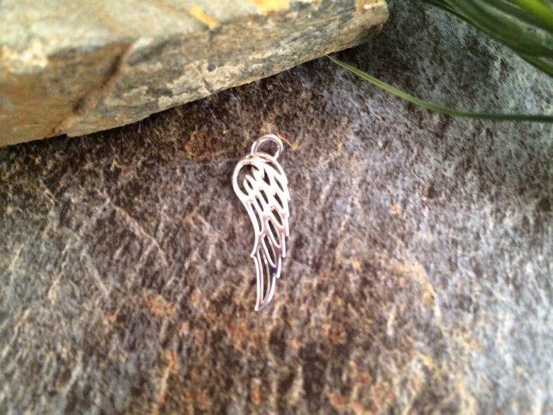 May include: A silver charm in the shape of an angel wing. The wing is detailed with intricate lines and curves.