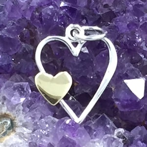 May include: A silver charm in the shape of a heart with a smaller gold heart inside. The charm is on a purple background.