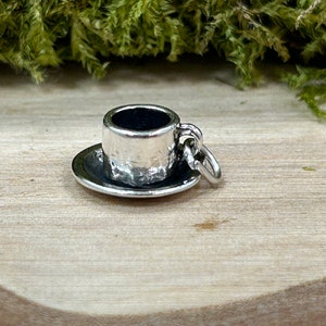May include: A silver charm in the shape of a small teacup and saucer. The charm has a textured surface and a loop for attaching to a bracelet or necklace.
