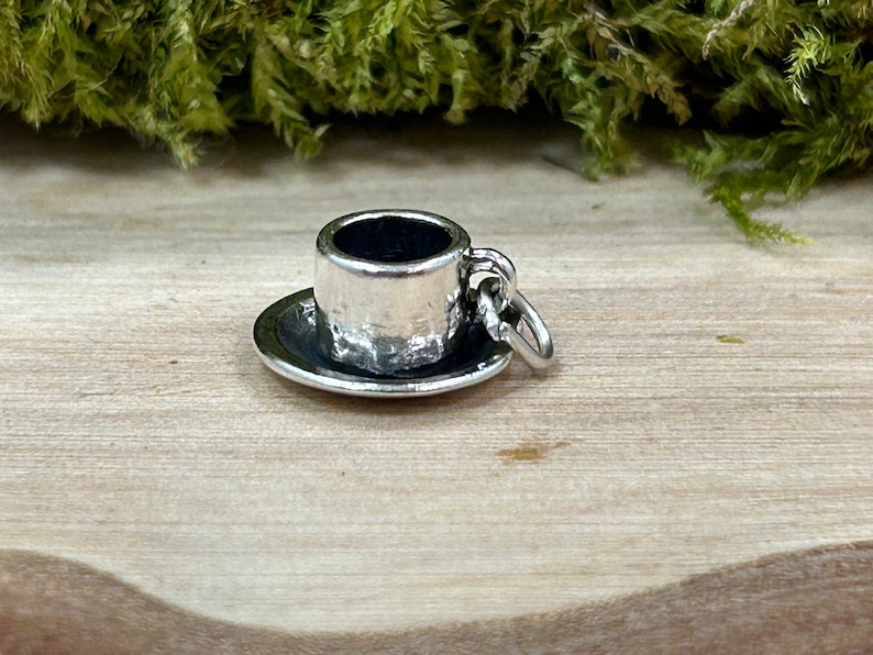 May include: A silver charm in the shape of a small teacup and saucer. The charm has a textured surface and a loop for attaching to a bracelet or necklace.