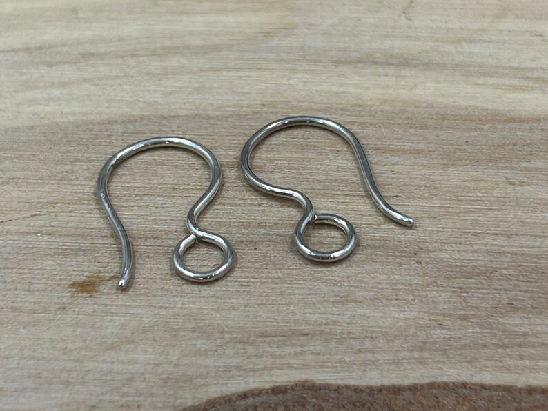 May include: Two silver earring hooks, also known as French hooks, are shown on a wooden surface. The hooks are made of metal and have a smooth finish.