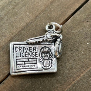May include: A silver charm featuring a driver's license design with the words "DRIVER LICENSE" and a smiling face. A key and a small decorative element are attached to the license. The charm is made of a shiny metal and is likely a pendant.