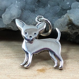 May include: A polished silver charm in the shape of a Chihuahua dog. The charm has a loop at the top for attaching to a bracelet or necklace. The dog is standing with its tail up and its ears perked.