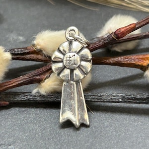 Puede incluir: Un charm de plata con forma de cinta, con el número "#1" en el centro. El charm tiene un lazo en la parte superior para sujetarlo a una cadena. El fondo incluye ramitas marrones y acentos esponjosos de color crema.