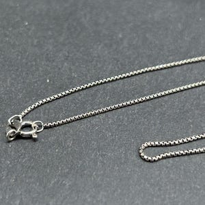 May include: A silver-colored necklace with a box chain design. The necklace features a small clasp at the end. The chain is made of small, square links, creating a delicate and elegant appearance. The necklace is displayed against a dark background.