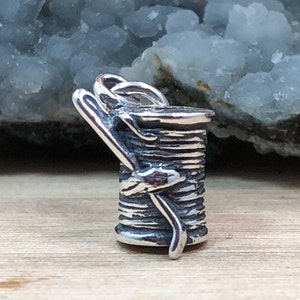May include: Silver charm in the shape of a spool of thread with a needle wrapped around it.