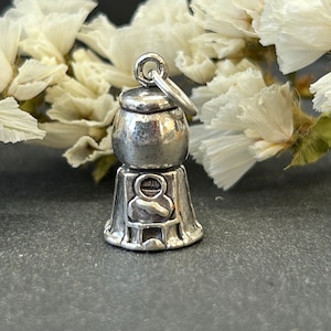 May include: Silver charm shaped like a gumball machine with a round top and a base with a person inside. The charm has a loop at the top for attaching to a bracelet or necklace.