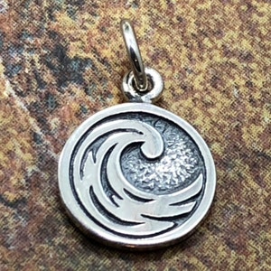 May include: Silver charm with a stylized wave design. The charm is round and has a textured surface.