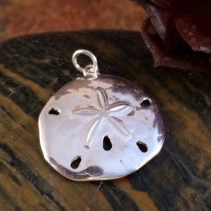 May include: A polished silver-toned pendant shaped like a sand dollar, featuring a central five-pointed star pattern and five small, elongated perforations arranged in a circular formation, topped with a small circular jump ring for attachment.