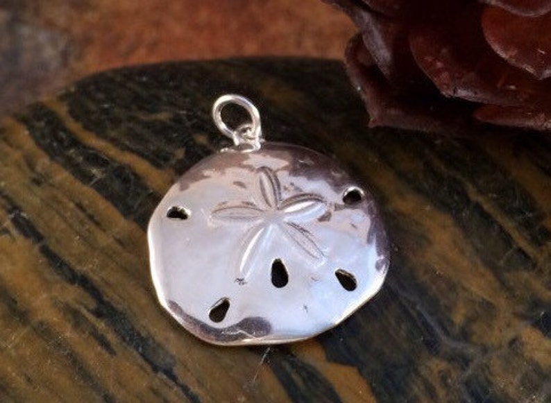 May include: A polished silver-toned pendant shaped like a sand dollar, featuring a central five-pointed star pattern and five small, elongated perforations arranged in a circular formation, topped with a small circular jump ring for attachment.