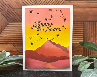 May include: A greeting card with a watercolor design. The card features a mountain landscape in shades of red and orange, with a yellow and orange sky. The text "Every journey starts with a dream" is written in black.