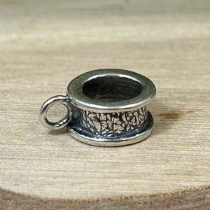 May include: Silver charm with a textured, cylindrical design and a small loop on the side. The charm is designed to be used as a bead on a bracelet or necklace.