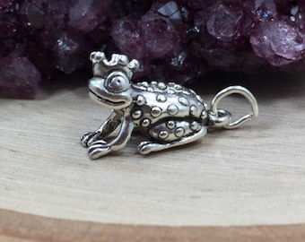 May include: A silver charm in the shape of a frog wearing a crown. The frog has a textured, bumpy surface and a small loop for attaching to a bracelet or necklace.