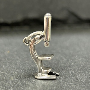 May include: A silver charm in the shape of a microscope. The microscope is detailed and has a round lens on top.