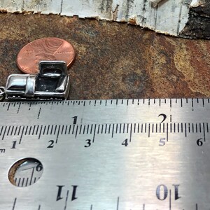May include: A small, silver-colored charm in the shape of a vintage pickup truck. The charm is approximately 0.75 inches long and has a detailed design. A ruler is placed next to the charm for scale, and a penny is also visible.