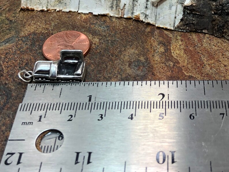 May include: A small, silver-colored charm in the shape of a vintage pickup truck. The charm is approximately 0.75 inches long and has a detailed design. A ruler is placed next to the charm for scale, and a penny is also visible.