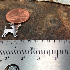 May include: A small, silver-colored pendant in the shape of a dog, likely a Chihuahua, next to a penny and a ruler. The pendant is approximately 1 inch long. The ruler shows measurements in inches. The background is a textured, brown surface.