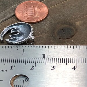 May include: A small, silver-colored pendant in a crescent moon shape, with a loop for a chain. A US one-cent coin is next to the pendant for size comparison. A ruler shows the pendant is approximately 0.75 inches long.