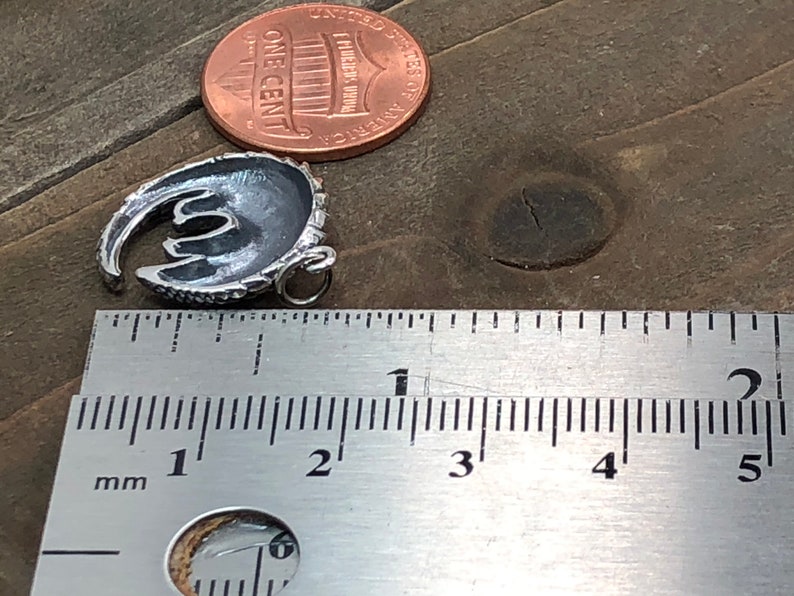 May include: A small, silver-colored pendant in a crescent moon shape, with a loop for a chain. A US one-cent coin is next to the pendant for size comparison. A ruler shows the pendant is approximately 0.75 inches long.